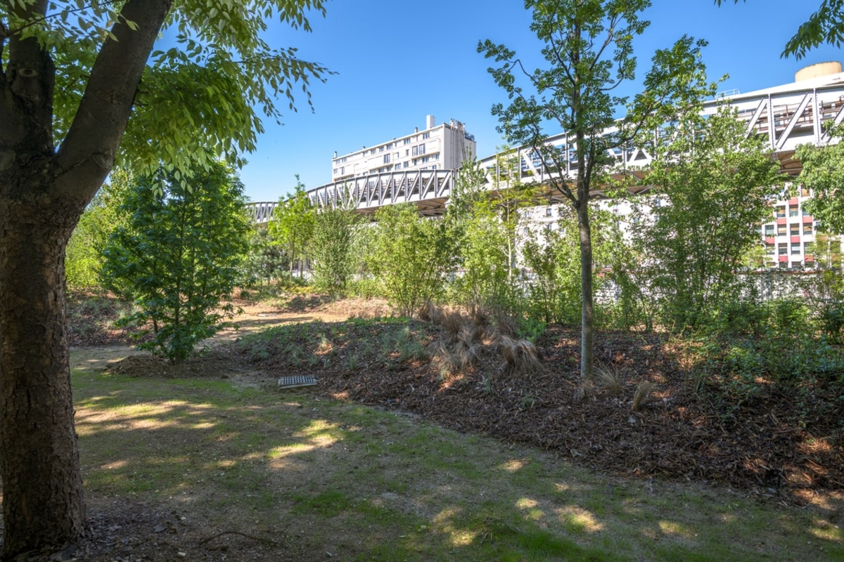 vue sur une partie du jardin, ombragée par des arbres nouvellement plantés, avec en arrière plan le métro aérien