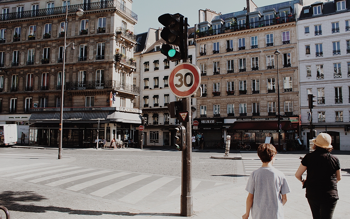 Deux personnes marchant en premier plan sur un trottoir, où est placé un feu tricolore auquel est fixé un panneau de limitation de vitesse à 30 km/h, dans une rue de Paris ensoleillée
