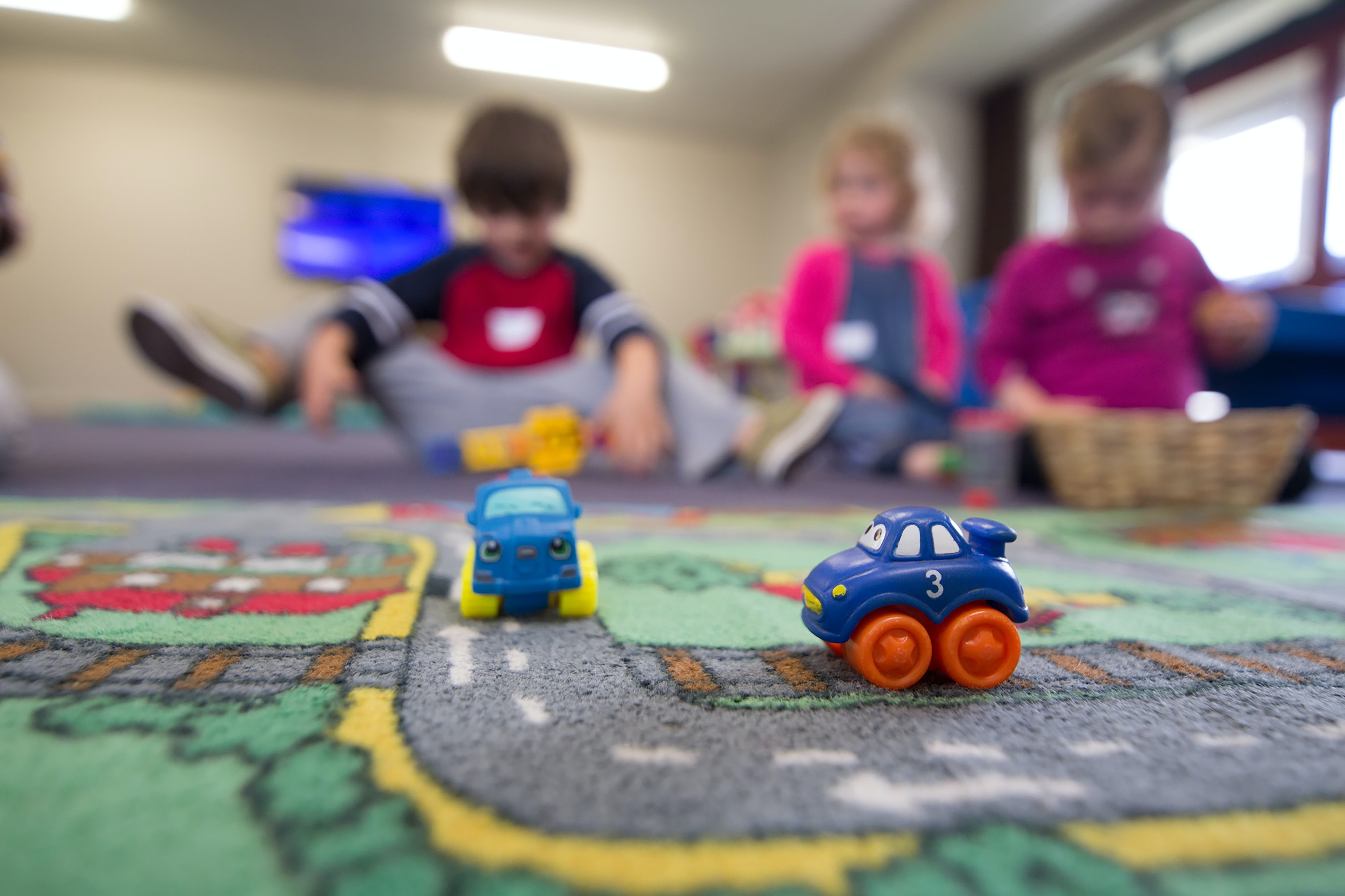 Deux petites voitures (jouets) en premier plan, 3 enfants flous au fond, dans une crèche