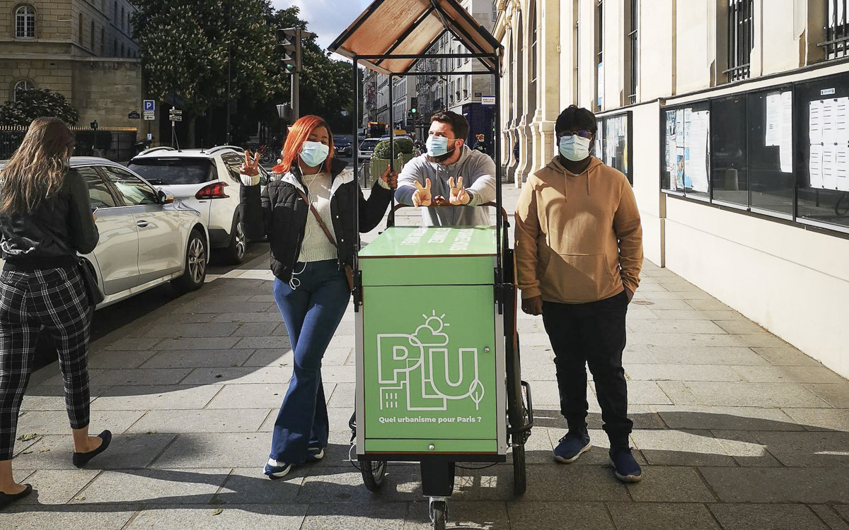 Triporteur vers avec le logo "PLU", entouré de 3 personnes amenées à animer les temps d'échange avec les habitants et habitantes.