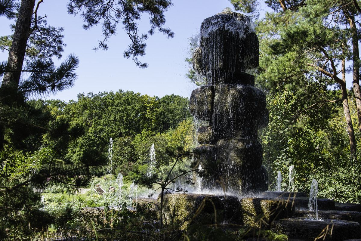 D'une sculpture en pierre monumentale jaillit des jets d'eau au côté d'un miroir d'eau, entouré d'arbres.