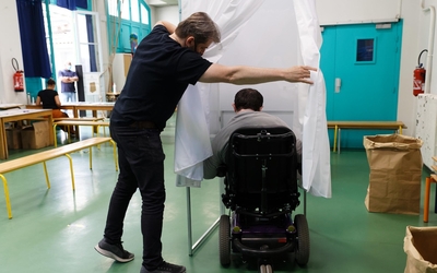 Un agent de la mairie aide un homme en fauteuil roulant à entrer dans un isoloir