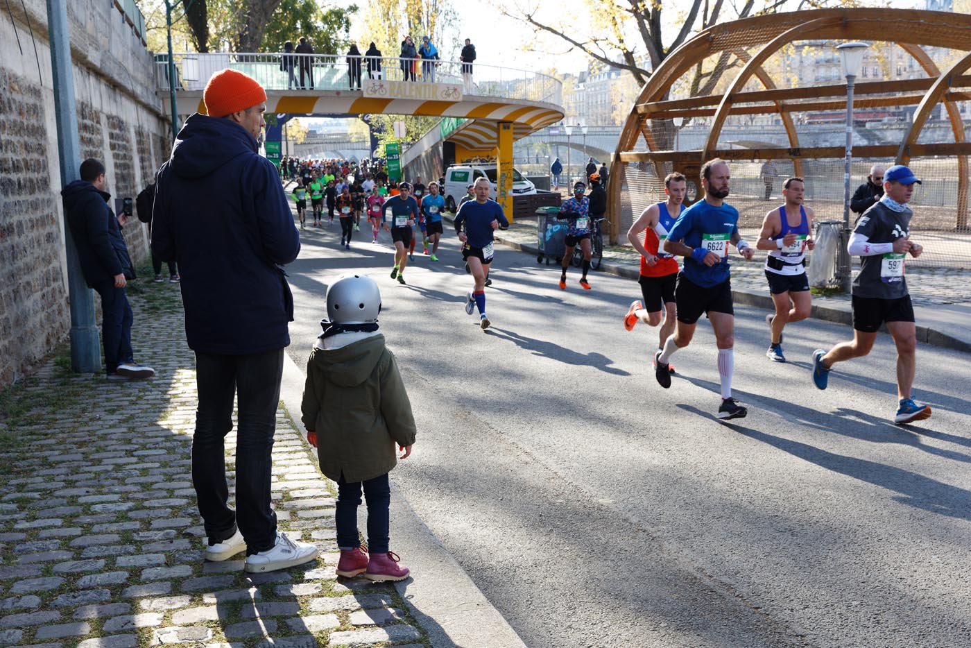 Un adulte et un enfant de dos, sur un trottoir, regardent passer des coureurs du marathon de Paris.