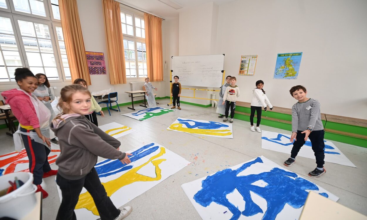 Des enfants montrent leurs peintures, de silhouettes colorées, sur de grandes feuilles de papier posées au sol