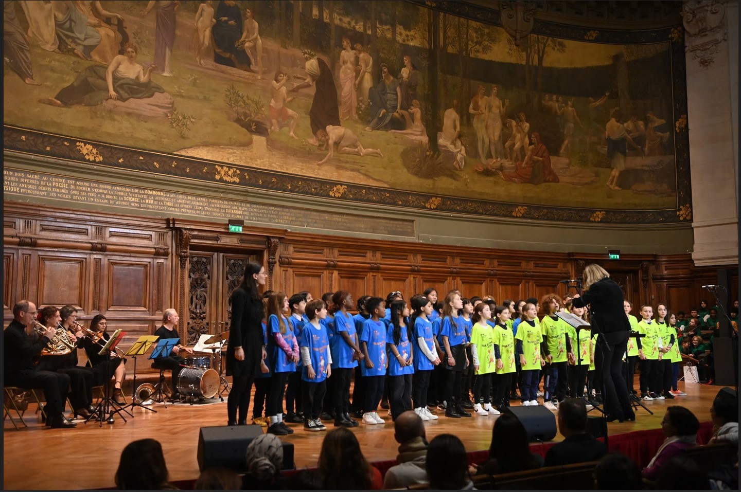 Sur l'estrade de l'amphithéâtre de la Sorbonne, les jeunes choristes en t-shirts de couleurs sont dirigés par la cheffe d'orchestre