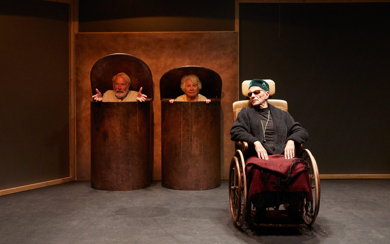 Sur la scène du Théâtre de l'Atelier, un personnage infirme devant deux personnes âgées placées dans des grandes poubelles
