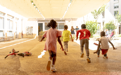 Des enfants courent sous le voile d'ombrage orange de la cour oasis de l'école Thionville, 19e