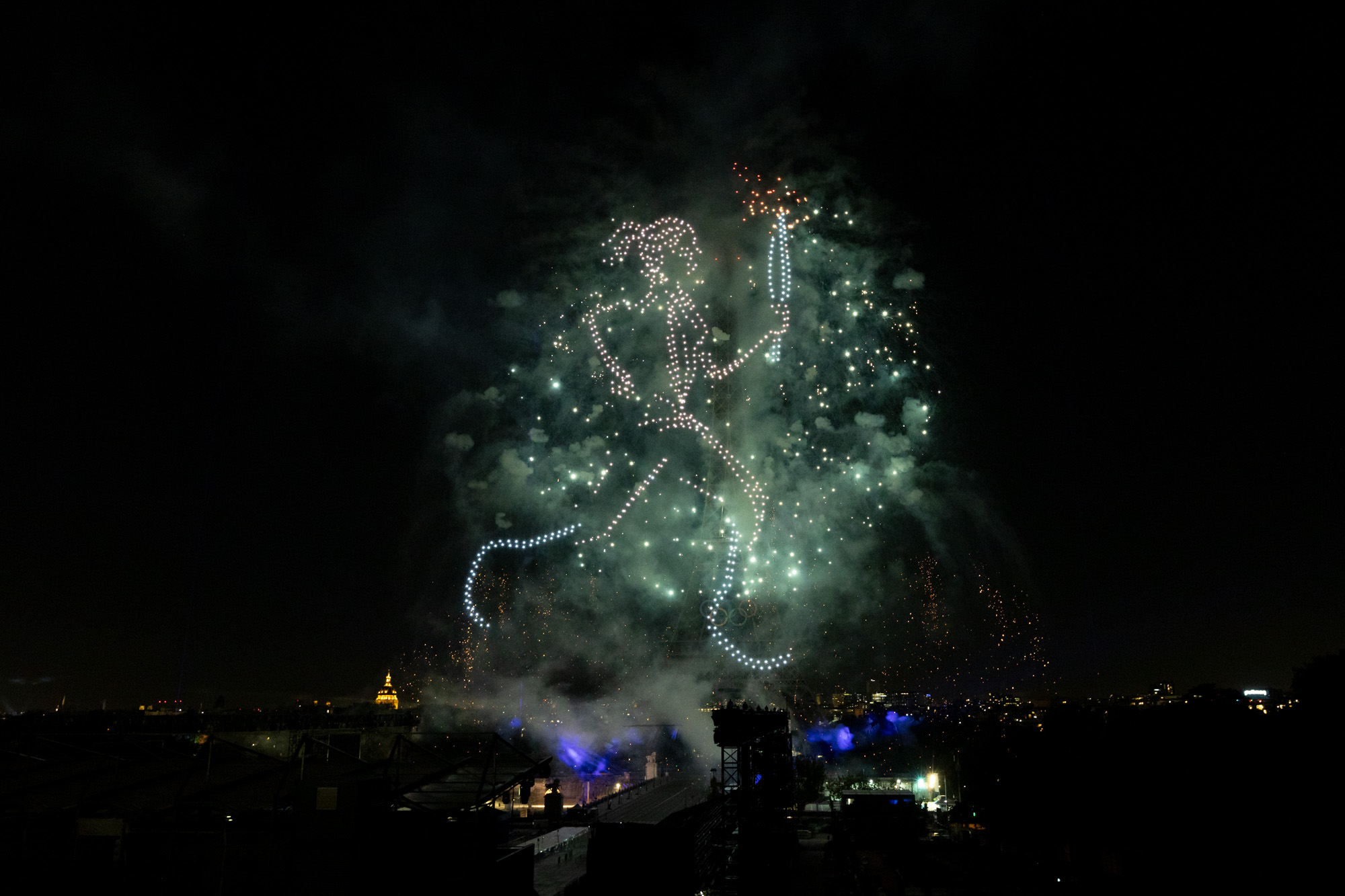 Un porteur de la flamme se dessine dans le ciel parisien grâce à l'usage de drones, face à la tour Eiffel.