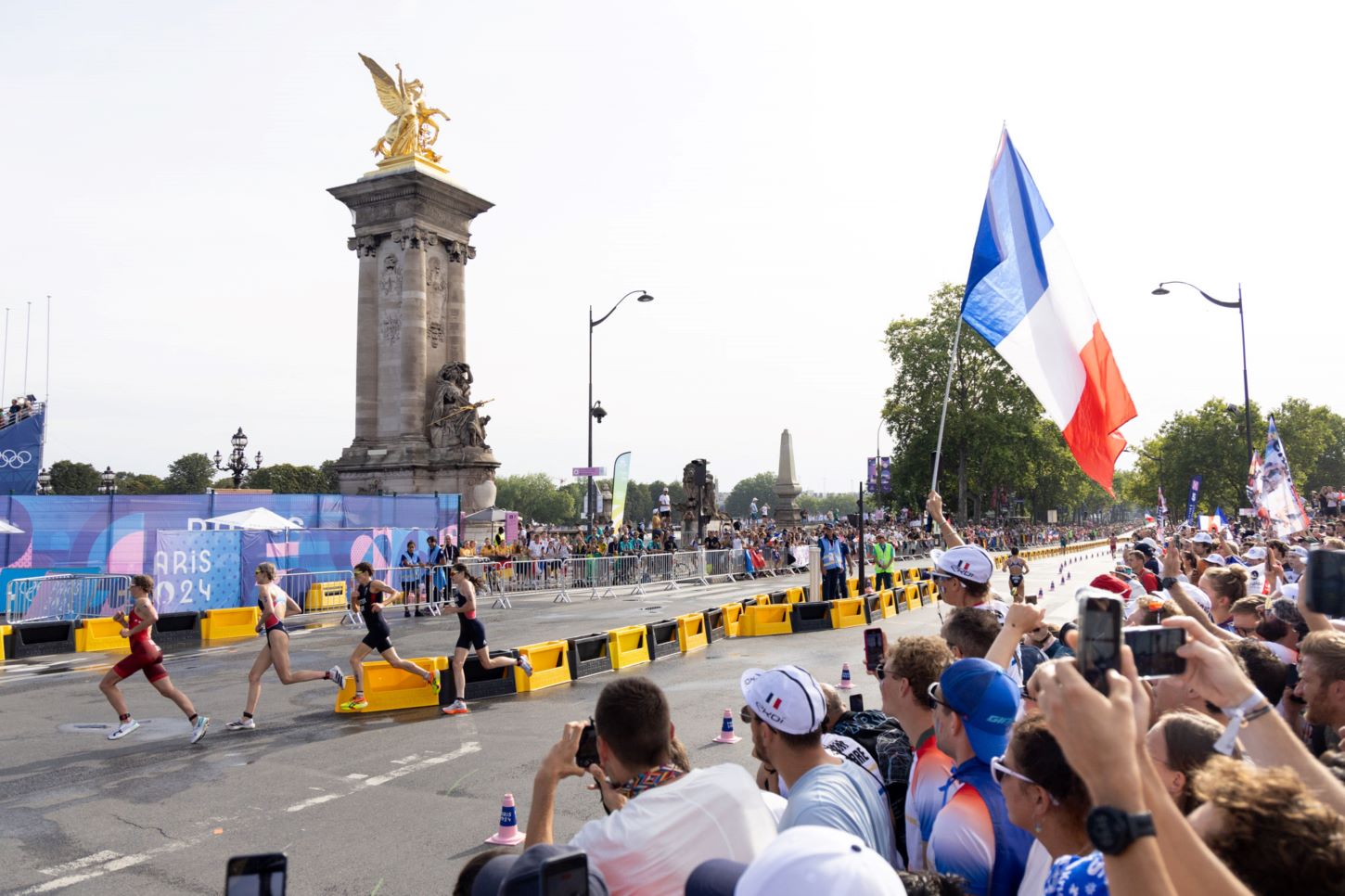 Une foule, dont l'un des membres brandit un drapeau français, assiste à l'épreuve de triathlon et voit quatre coureuses passer devant elle.