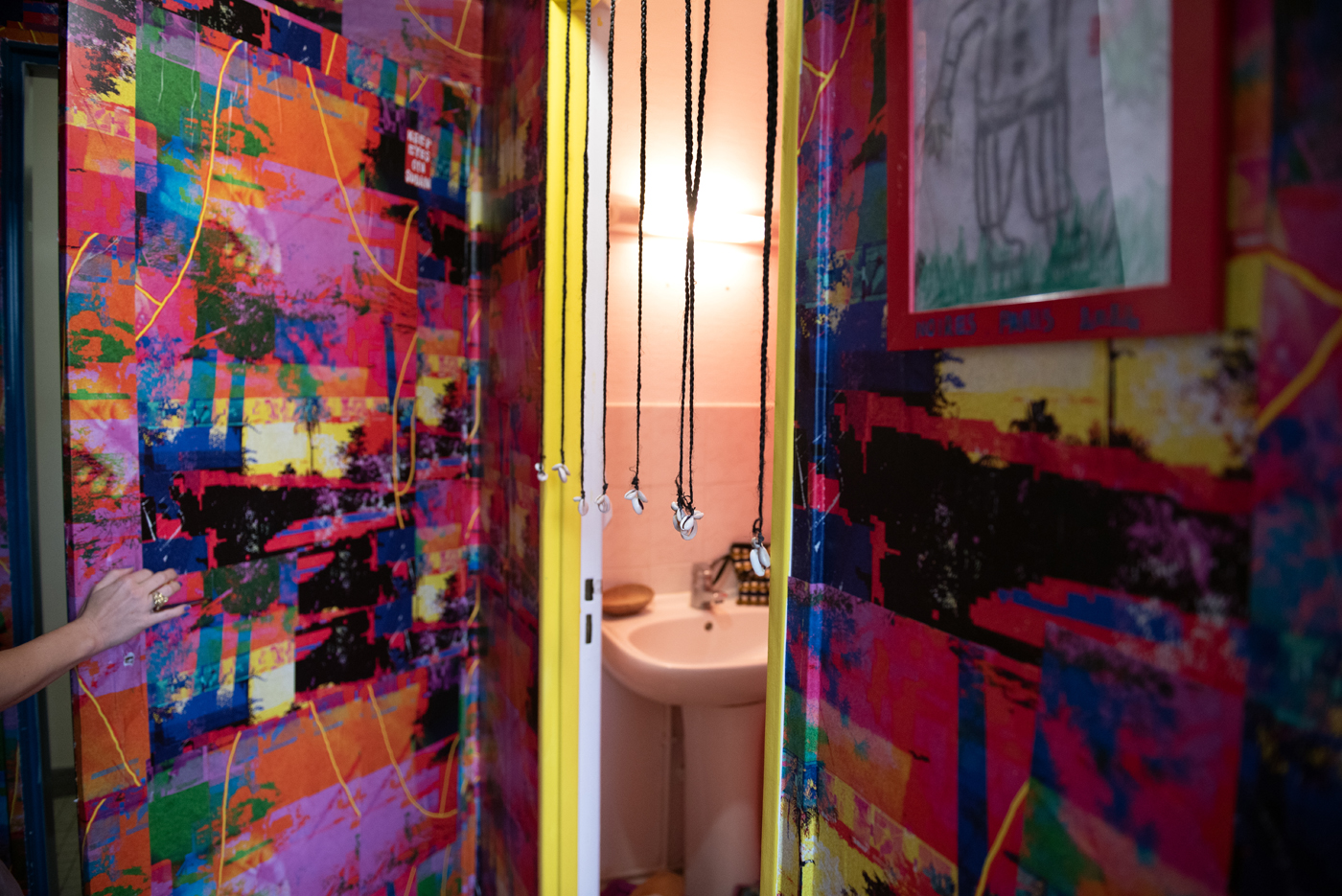 L'image montre l'entrée d'une salle de bain à travers une porte encadrée de jaune vif. Les murs autour sont couverts de peintures explosives aux couleurs vives - rose, bleu, vert, orange - créant une mosaïque abstraite. Des pendentifs sur des cordons noirs pendent devant la porte. À l'intérieur, on aperçoit un lavabo blanc simple. Une main apparaît sur la gauche, touchant le mur coloré.