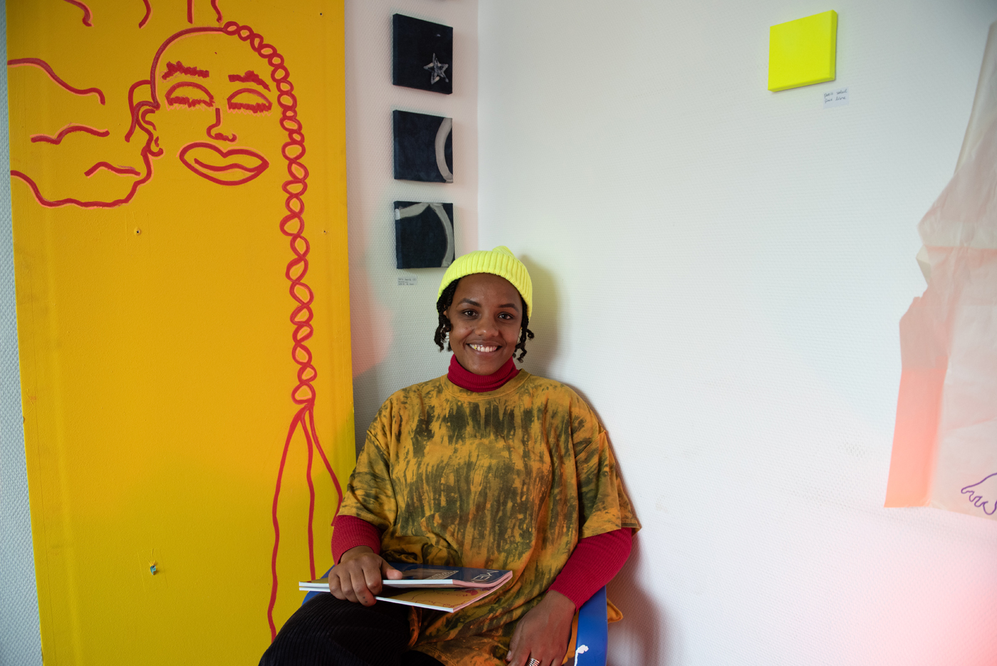 Une personne souriante est assise devant un mur décoré d'une grande fresque jaune avec un visage dessiné en rouge. Elle porte un bonnet jaune fluo, un t-shirt tie-dye jaune et marron, et un col roulé rouge en dessous. Elle tient des cahiers dans ses mains. Au mur, trois petits tableaux noirs sont alignés verticalement. L'ambiance est artistique et joyeuse.