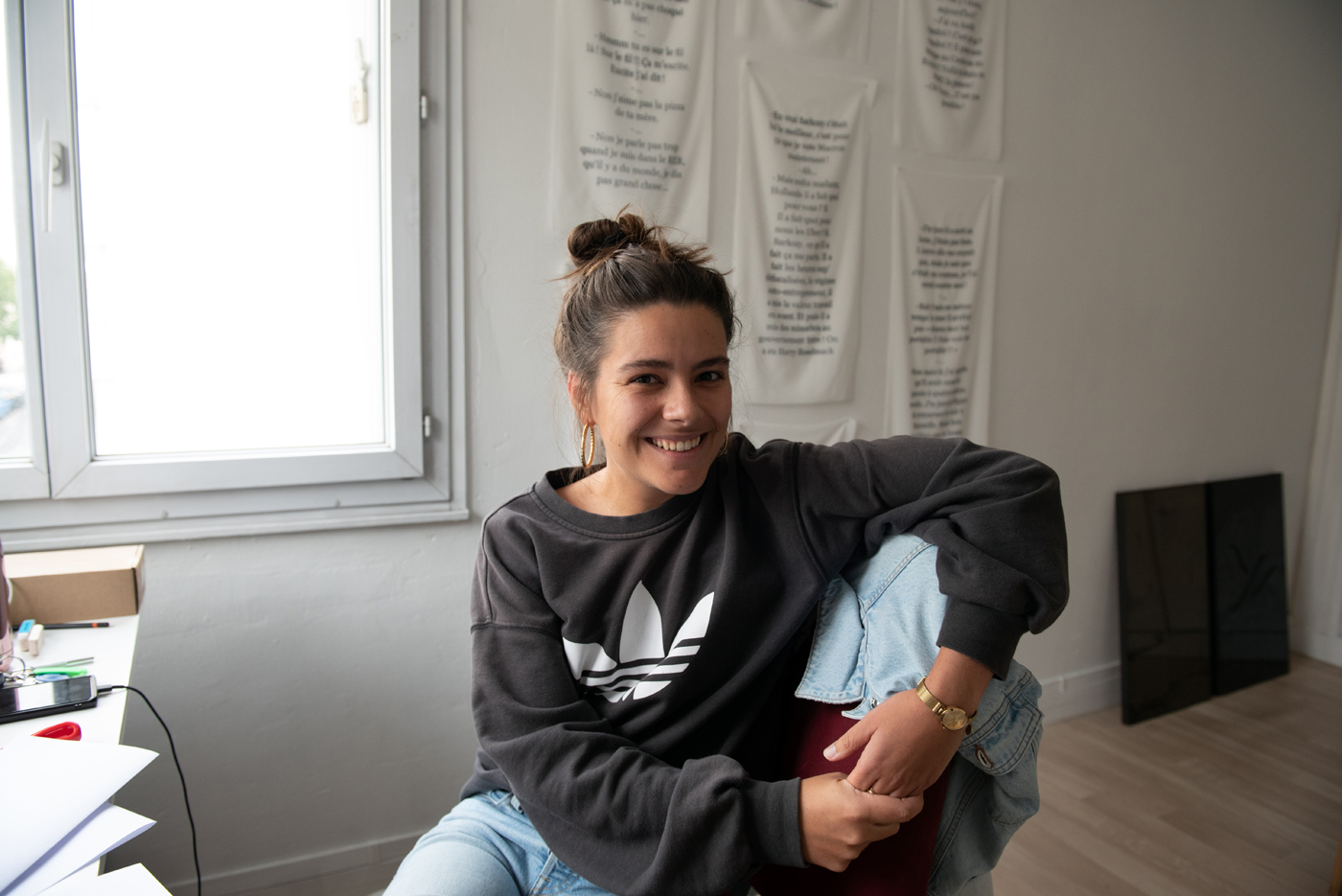 Dans une pièce lumineuse aux murs blancs, une personne souriante est assise de façon décontractée. Elle porte un sweat-shirt noir Adidas et un jean clair, ses cheveux sont relevés en chignon. Des textes sont affichés au mur derrière elle. La lumière naturelle entre par une fenêtre à gauche. L'ambiance est chaleureuse et détendue, comme dans un bureau ou un espace créatif personnel.