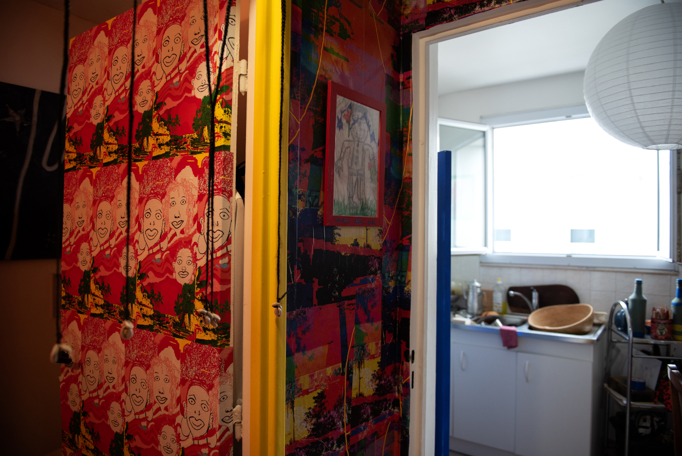 Une vue d'un espace artistique coloré montrant l'entrée d'une cuisine. Au premier plan, un mur est couvert d'un motif répétitif de visages souriants dessinés sur fond rouge. L'encadrement de porte est peint en jaune vif, et les murs adjacents sont décorés de peintures multicolores. À travers la porte, on aperçoit une cuisine blanche simple avec un évier et quelques ustensiles. Une lampe boule blanche est visible à droite.