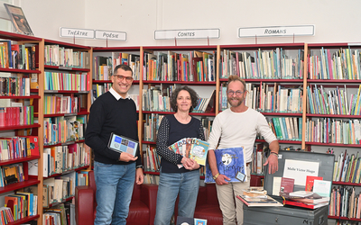 Trois animateurs du Centre Paris Lecture posent devant une bibliothèque avec des ouvrages, une tablette numérique et un carnet de dessin dans leurs mains