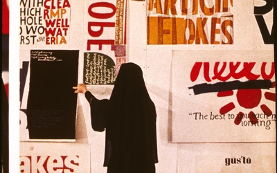 Image représentant une femme de dos vêtue d'un habit religieux noir face à un mur rempli d'affiches