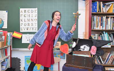 Sabrina Perez, animatrice lecture sourit, dans une classe sur fond de tableau et de livres avec une baguette magique dans la main