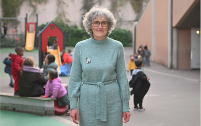 Nadine, animatrice en école maternelle pose dans la cour de son école sur fond d'enfants qui jouent autour d'un tobogan 