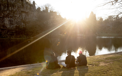 Des personnes sont assises sur l'herbe et se reposent au bord du lac du parc des Buttes Chaumont.
