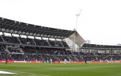 Stade de football avec tribunes remplies de supporters et joueurs sur le terrain au premier plan.