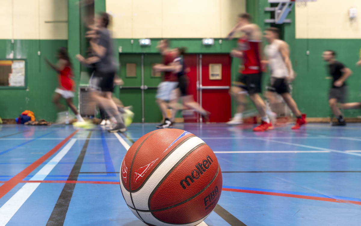 Personnes en arrière-plan s'entraînant dans un gymnase avec un ballon de basket au premier plan. 