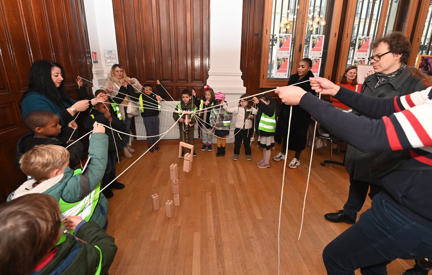 Des petits enfants et des adultes jouent à un jeu construction collaboratif  ou il s'agit d'empiler des cubes de bois en manipulant des ficelles