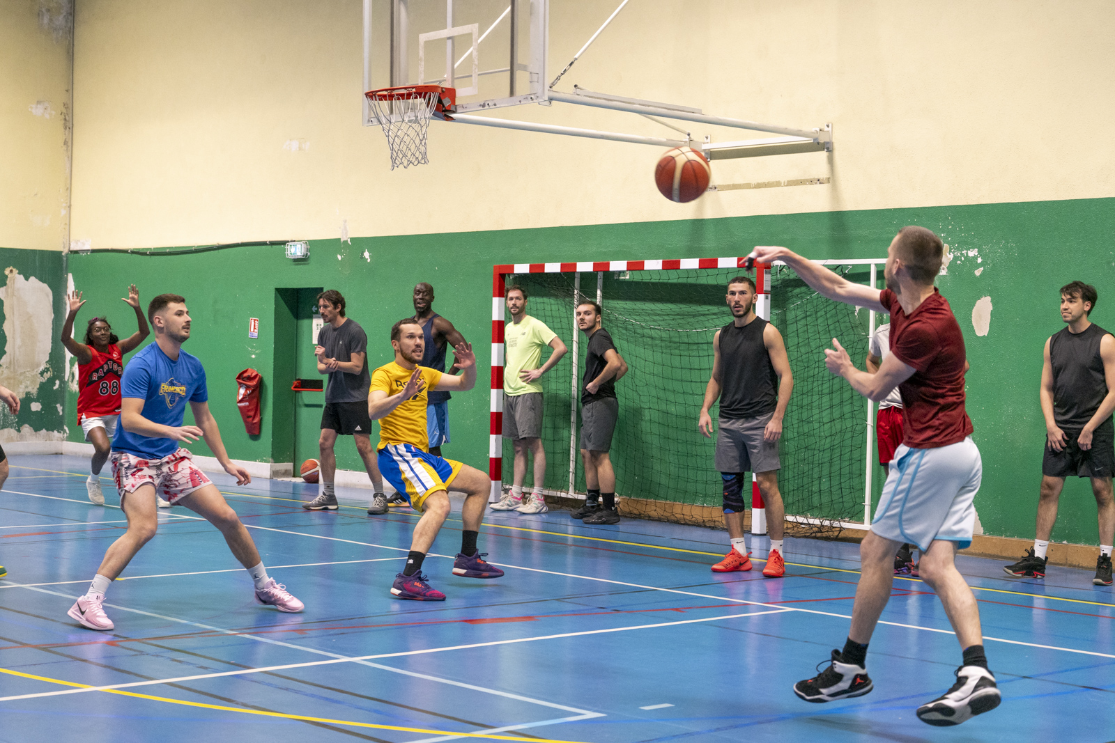 Des joueurs de basket se font des passes sous un panier dans un gymnase au sol bleu.