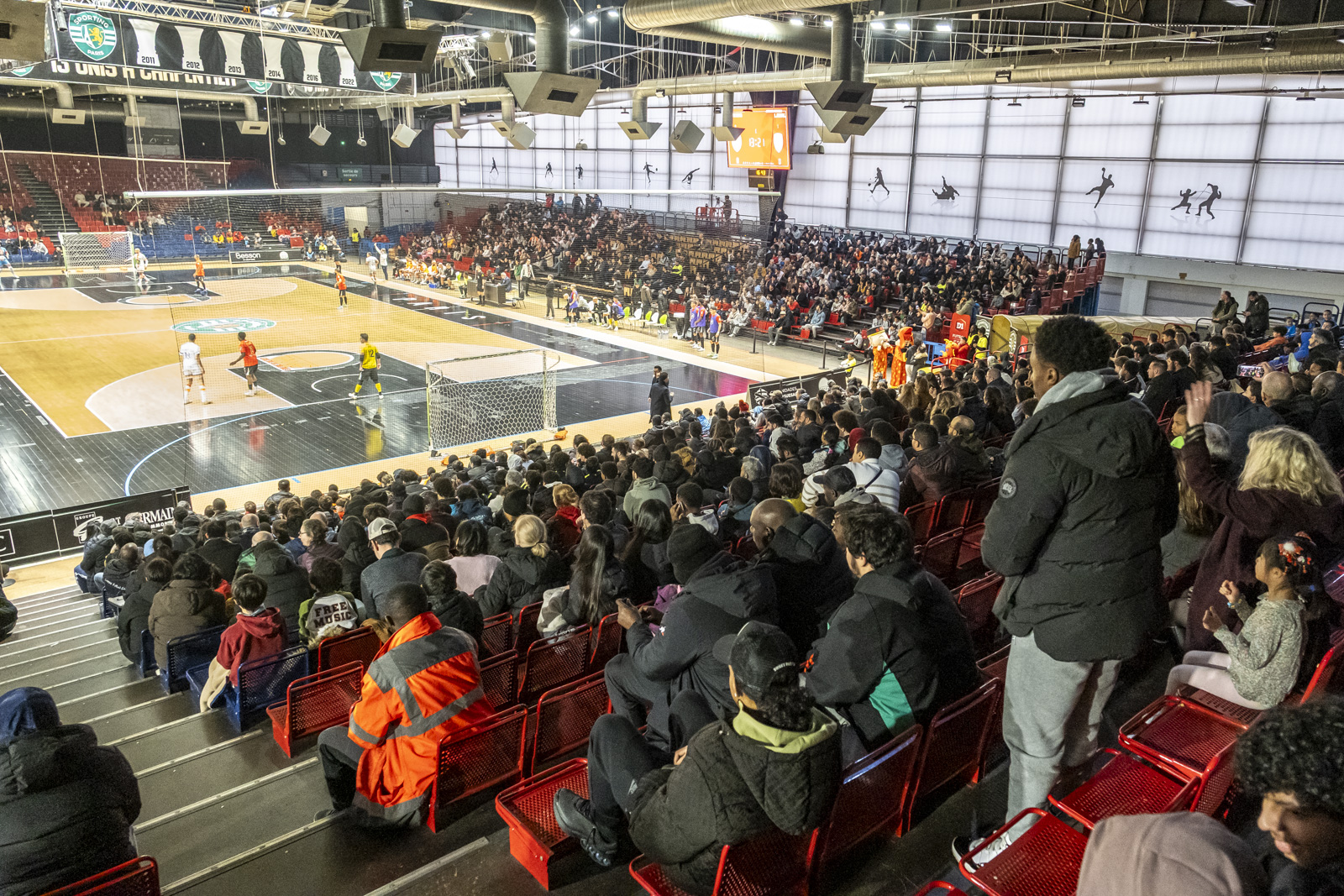 Des gens regardent un match de futsal depuis les tribunes d'une salle couverte.