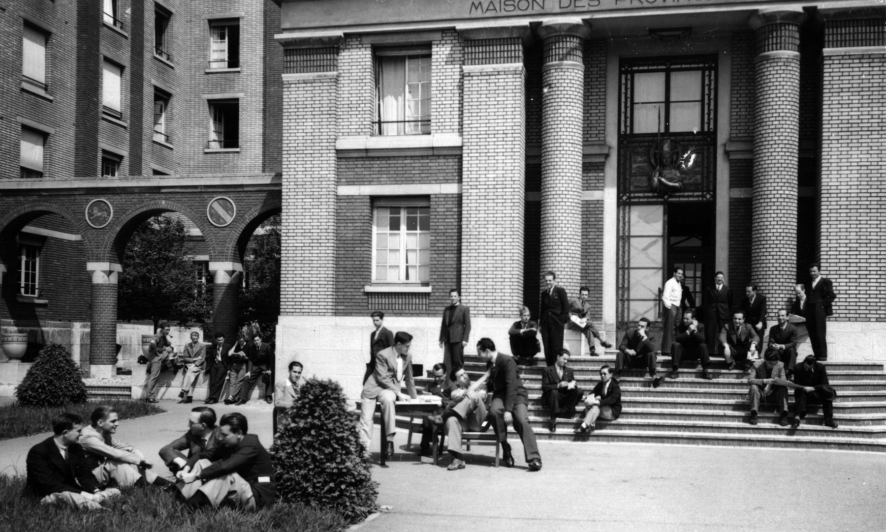 Une trentaine de jeunes hommes en costume devant un bâtiment. Photo noir et blanc