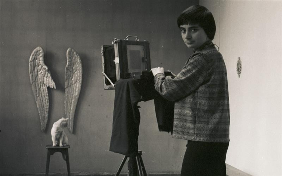 Autoportrait en noir et blanc d'Agnès Varda debout tenant sa cemara. Des ailes d'anges sont suspendu sur le mur et sa chatte nini sur un banc