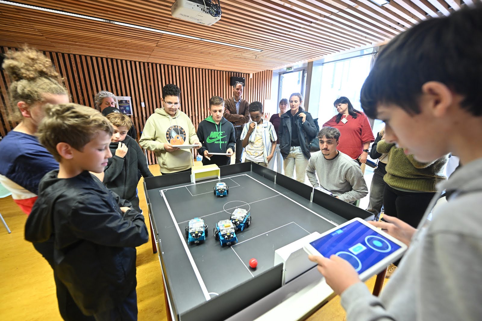 4 collégiens dirigent à l'aide tablette les 4 robots présents sur le terrain de foot et un large groupe de supporter sr former autour d'eux