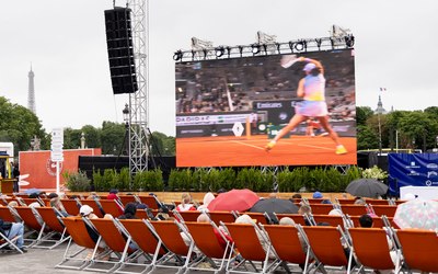 Rangées de transats avec des personnes assises qui ont parfois un parapluie pour suivre un match de tennis sur écran géant