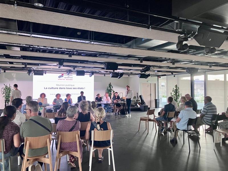 Photo prise en début de réunion depuis le fond de la salle, où l'on peut voir  le public de dos et les intervenants à la tribune