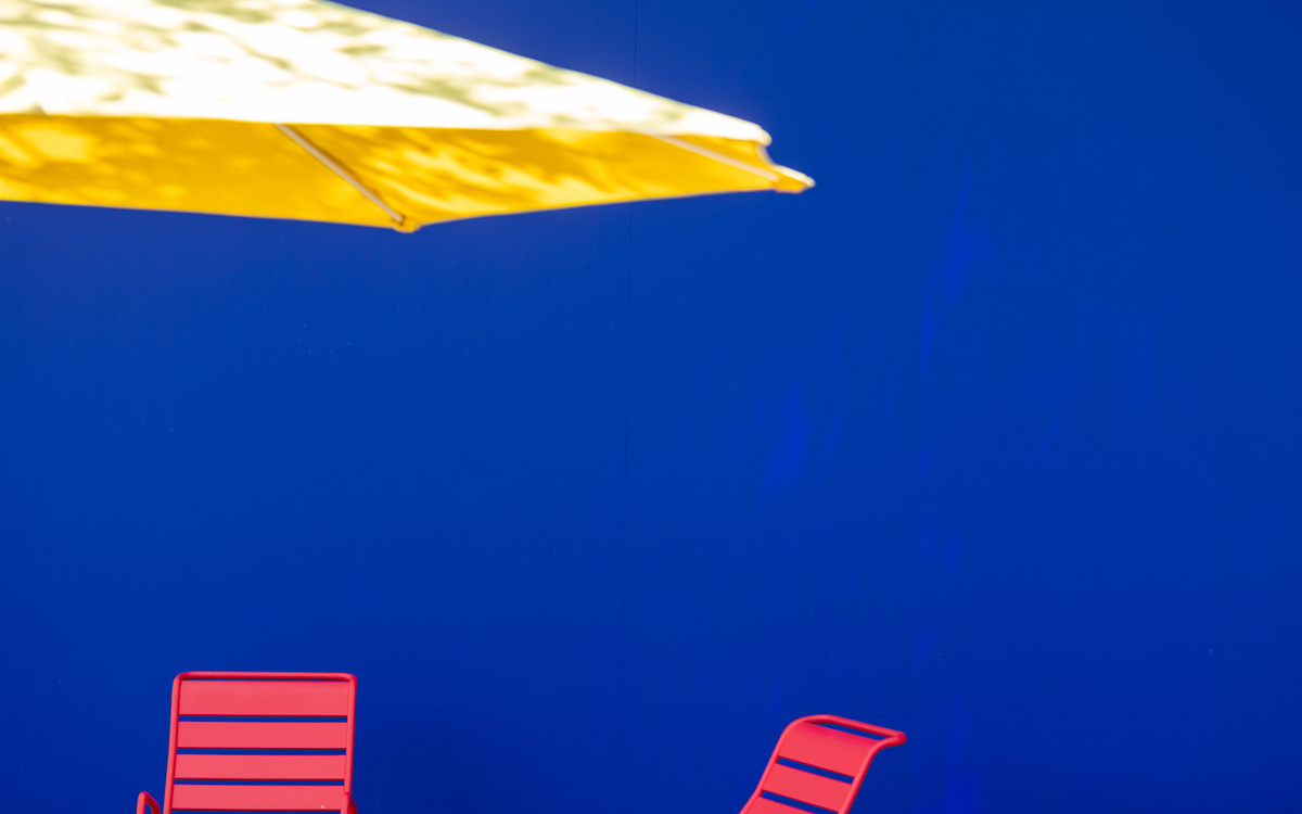 Image très graphique de Paris plage, représentant un mur bleu, un parasol jaune à gauche et des dossiers de chaises roses.