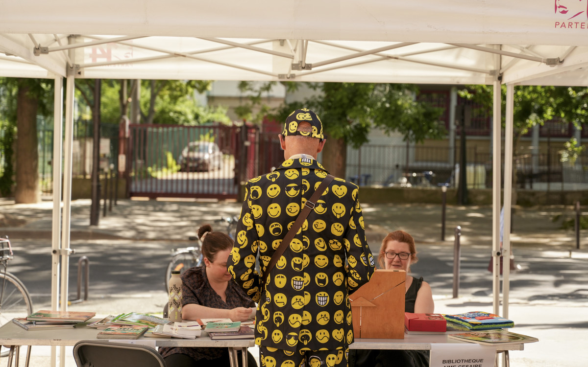 Monsieur debout de dos avec une veste à motifs smiley s'adressant à deux représentantes d'une association assises sous un barnum