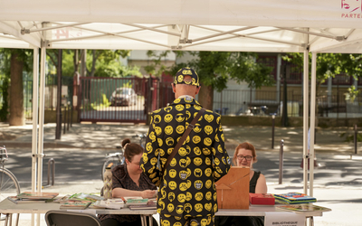 Monsieur debout de dos avec une veste à motifs smiley s'adressant à deux représentantes d'une association assises sous un barnum