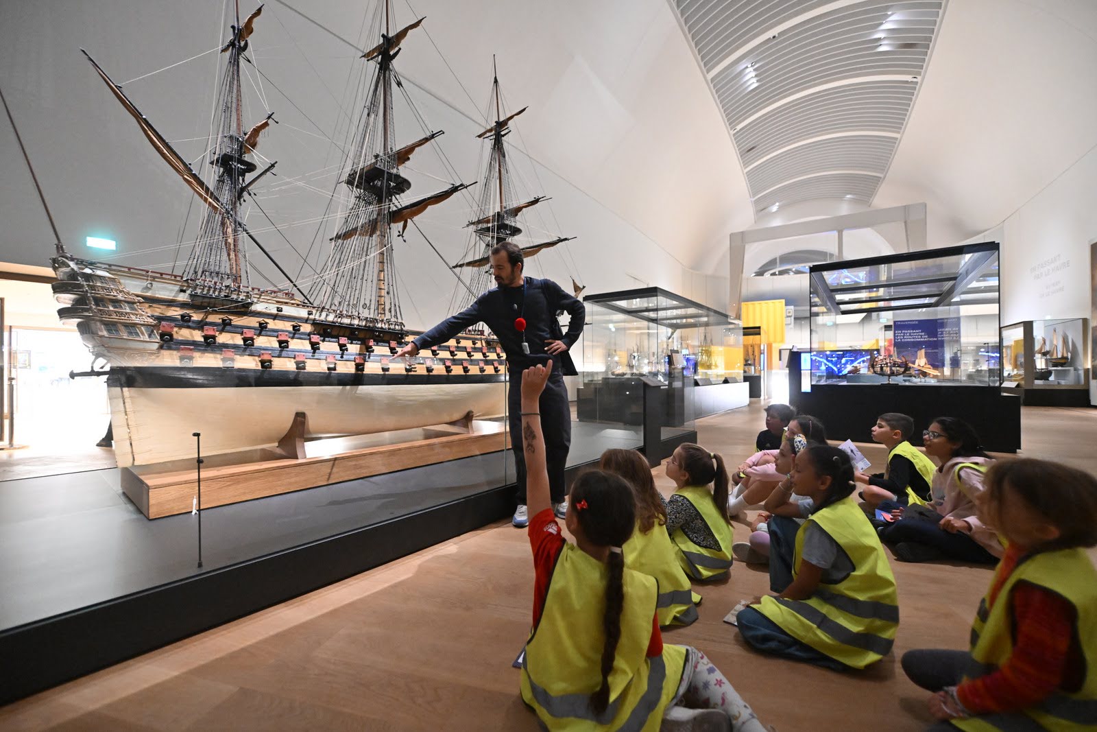 Les enfants écoutent les explications assis face à une maquette de voilier