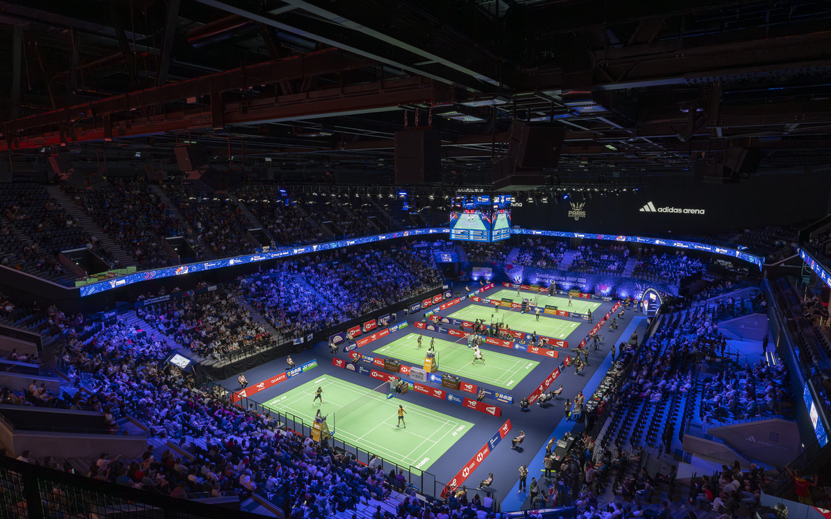 Vue aérienne d’une arène remplie de spectateurs lors d’un tournoi de badminton. Quatre matchs se déroulent simultanément sur des terrains verts éclairés par des projecteurs, entourés de publicités colorées.