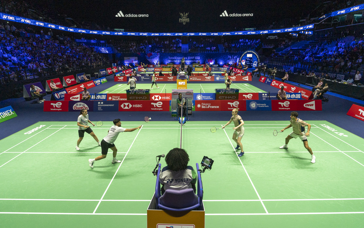 Match de double en badminton dans une arène remplie de spectateurs. Quatre joueurs, deux par équipe, s'affrontent sur un terrain vert éclairé. Un arbitre est assis sur une chaise surélevée au premier plan.