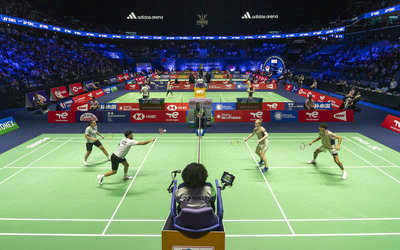 Match de double en badminton dans une arène remplie de spectateurs. Quatre joueurs, deux par équipe, s'affrontent sur un terrain vert éclairé. Un arbitre est assis sur une chaise surélevée au premier plan.