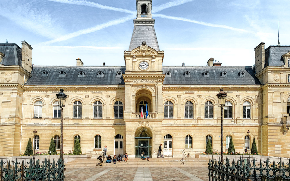 Voici une description adaptée pour le texte alternatif (attribut "alt") d’un site web : **"Façade d’un bâtiment historique en pierre claire avec une horloge centrale, un clocher et des drapeaux français et européen. L’architecture est typique des mairies ou institutions publiques, avec des fenêtres symétriques et un escalier menant à une entrée monumentale. Des personnes sont assises sur les marches et des lampadaires ornent la cour pavée devant le bâtiment