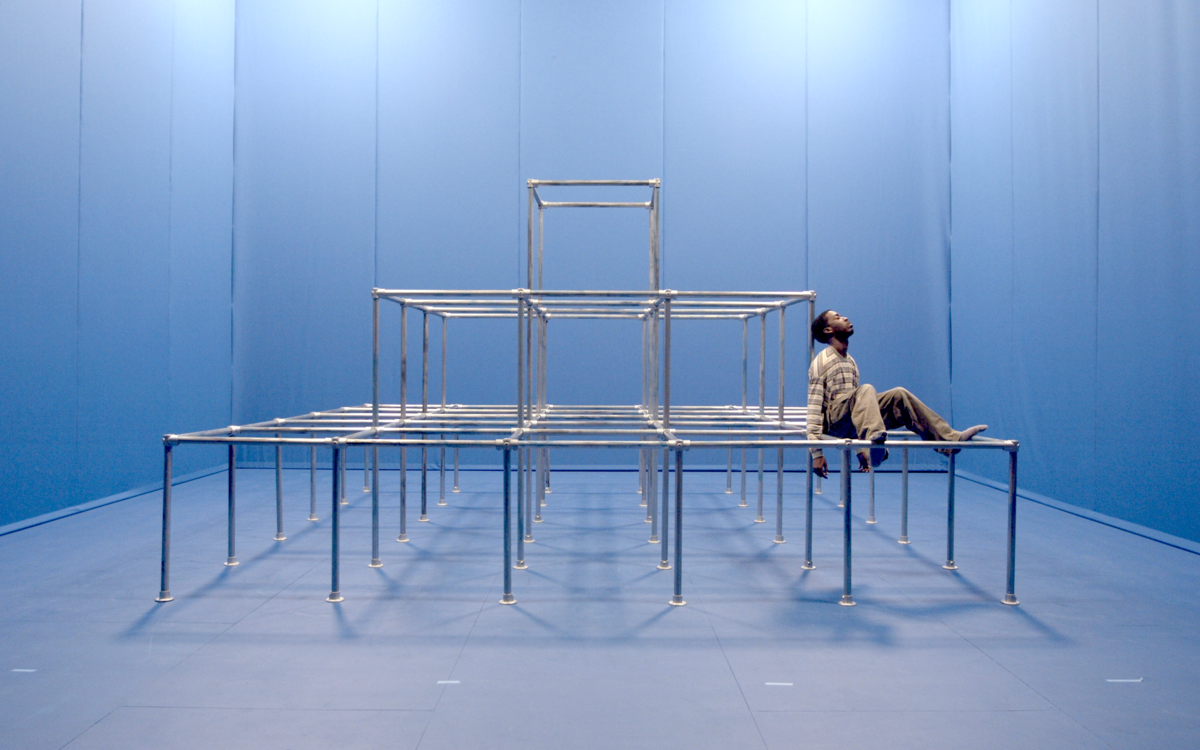 Dans une pièce bleu, un homme assis sur une sculpture métallique graphique