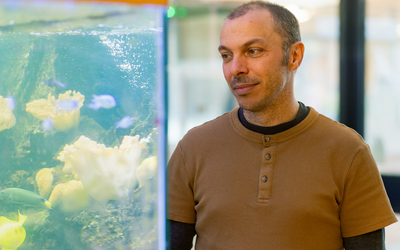 Un homme portant un t-shirt henley marron à manches courtes sur un t-shirt noir à manches longues se tient près d'un aquarium et regarde vers la droite avec une expression pensive. L'aquarium à sa gauche contient plusieurs méduses jaunes et blanches flottant dans l'eau turquoise illuminée. La surface de l'eau est visible en haut de l'aquarium avec des reflets lumineux. L'arrière-plan montre un espace intérieur flou avec des éléments architecturaux et de l'éclairage naturel. La composition capture un moment de contemplation dans un environnement aquatique éducatif.