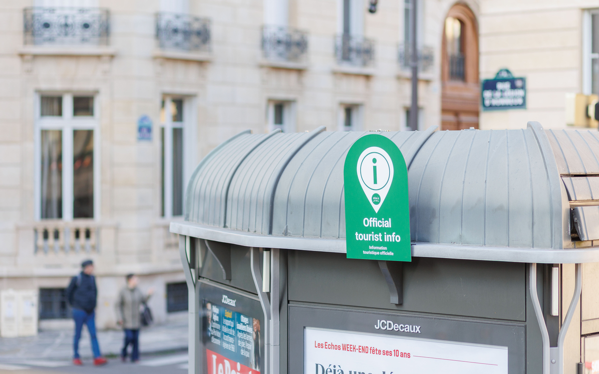 Sur la photo, on aperçoit l’un des 37 kiosques participant au dispositif tourisme : devant, une petite pancarte colorée indique qu’il fait partie du réseau, signalant ainsi aux passants qu’ils peuvent y trouver des conseils pour découvrir Paris.