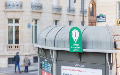 Sur la photo, on aperçoit l’un des 37 kiosques participant au dispositif tourisme : devant, une petite pancarte colorée indique qu’il fait partie du réseau, signalant ainsi aux passants qu’ils peuvent y trouver des conseils pour découvrir Paris.