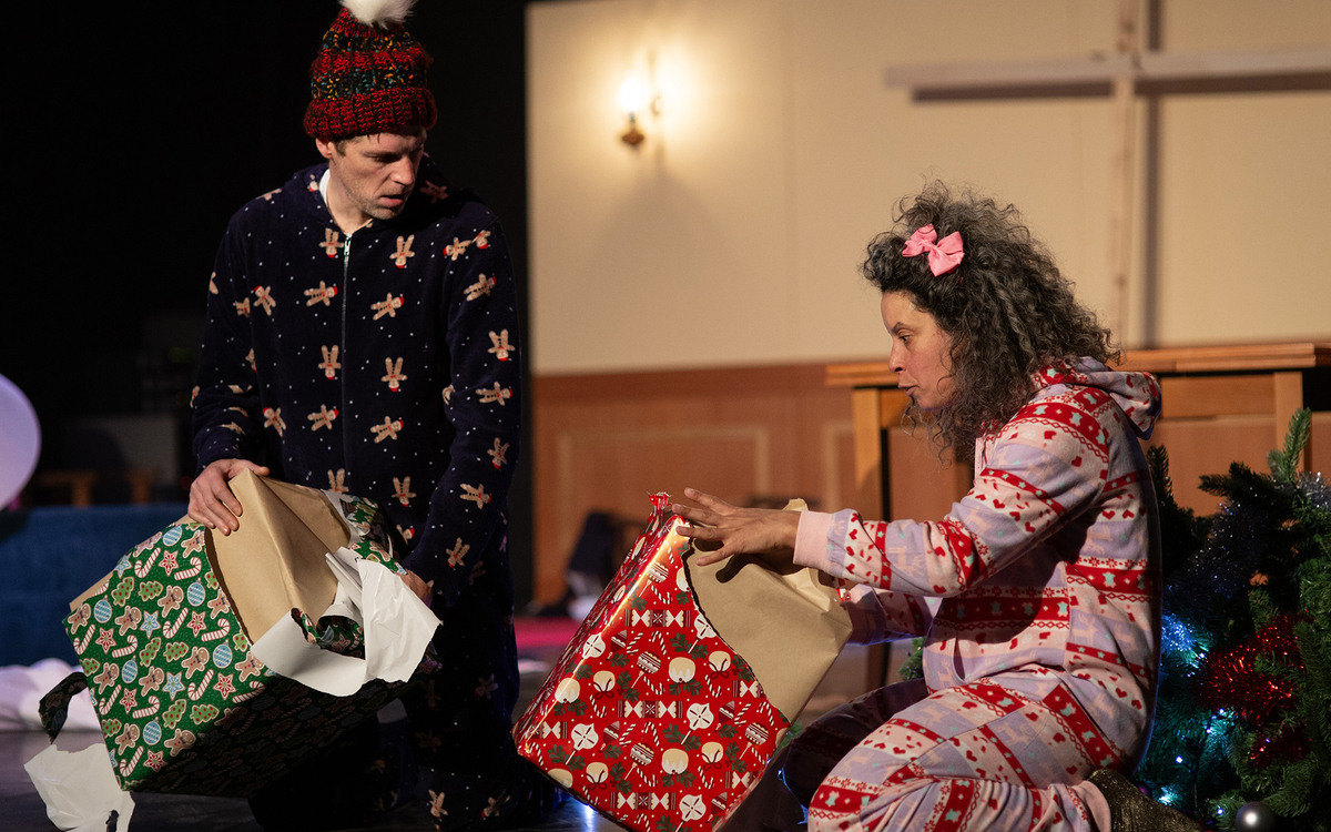 Sur une scène de théâtre, un homme et une femme glissés dans des pyjamas de Noël déballent des cadeaux