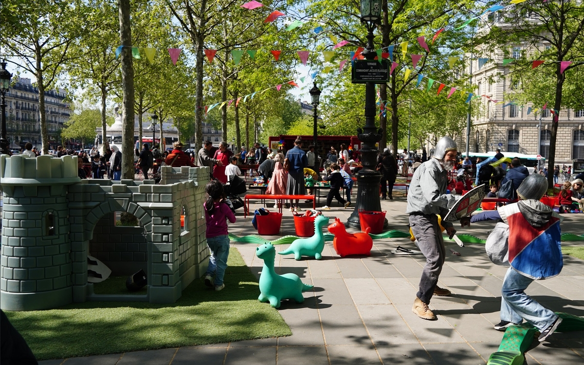 Espace scénographié sur la place de la République à Paris avec un château-fort pour enfants à gauche, à droite un adulte et un enfant déguisés en chevaliers dans un combat à l'épée, des petits dinosaures au sol, des jeux divers joués sur des tables en arrière plan.