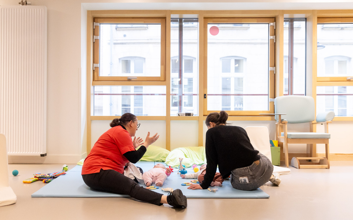 Deux femmes sont sur un tapis avec deux bébés allongés sur les dos. Elles jouent avec eux. 