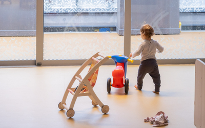 Vue d'un enfant de dos, à côté d'un objet roulant. En arrière plan, une terrasse derrière une grande baie vitrée. 