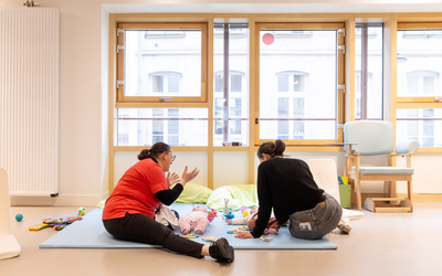 Deux femmes sont sur un tapis avec deux bébés allongés sur les dos. Elles jouent avec eux. 