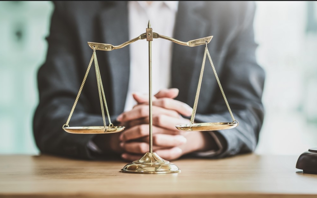 Photo d'un avocat assis à son bureau, joignant ses mains entre elles. Devant lui se trouve une balance (symbole de la justice) et un marteau